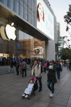 China, Shanghai, Modern young Chinese family with daughter in stroller and infant in sling stroll past the Apple store on Nanjing Lu, Store staff and Security on steps at entrance, evening light.