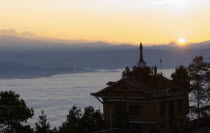 Nepal, Nagarkot, Sunrise view across clouded valley towards Himalayan mountains with Buddhist Stupa on top of building.