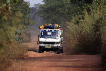 Gambia, Minibus driving down narrow dirt road covered in red soil, carrying provisions on roof rack.