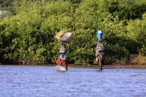 Gambia, Two women crossing water in bare feet carrying walking stick and rice in bucket and straw on their heads. 