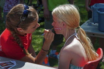 Canada, Alberta, Lethbridge, A bare-shouldered young blonde Canadian girl sits to have the word CANADA painted on her face at Canada Day celebrations at Henderson Park, Face painter in a red T shirt h...