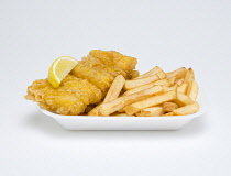 Food, Cooked, Fish, A portion of battered deep fried huss with a slice of lemon and potato chips in an insulated polystyrene foam tray on a white background.