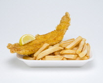 Food, Cooked, Fish, A portion of battered deep fried cod with a slice of lemon and potato chips in an insulated polystyrene foam tray on a white background.