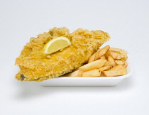 Food, Cooked, Fish, A portion of battered deep fried plaice with a slice of lemon and potato chips in an insulated polystyrene foam tray on a white background.