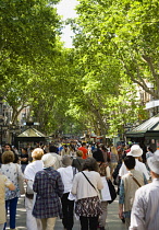 People walking along the central tree shaded walkway of La Rambla historic avenue past stalls in the Old Town district.