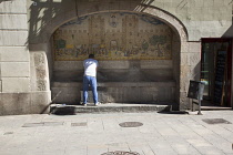 Spain, Catalonia, Barcelona, Man washing hands in drinking water fountain in Portaferrissa Street off La Rambla in the Gothic Quarter with a tiled depiction of one of the gates to the old walled city.