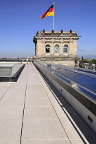 Germany, Berlin, German flag fluttering on a corner tower of the Reichstag building as seen from the rooftop terrace with two tourists strolling.