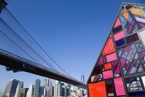 USA, New York, Brooklyn Bridge Park, colourful plexiglass house called Kolonihavehus by sculptor Tom Fruin below the suspension bridge spanning the East River with Lower Manhattan beyond.