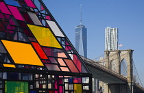 USA, New York, Brooklyn Bridge Park, colourful plexiglass house called Kolonihavehus by sculptor Tom Fruin below the suspension bridge spanning the East River with Lower Manhattan beyond.