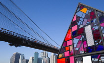 USA, New York, Brooklyn Bridge Park, colourful plexiglass house called Kolonihavehus by sculptor Tom Fruin below the suspension bridge spanning the East River with Lower Manhattan beyond.