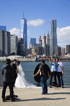 USA, New York, Brooklyn Bridge Park, two young men taking a selfie with the Lower Manhattan skyscraper skyline as a backdrop beside a couple having their wedding photograph taken on the Fulton Ferry L...