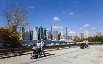USA, New York, Brooklyn Bridge Park, couple in autumn pushing prams along a path between Pier 1 and Pier 2 with the Lower Manhattan skyscraper skyline and the East River beyond.