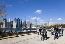 USA, New York, Brooklyn Bridge Park, walkers and cyclists on a path in autumn between Pier 1 and Pier 2 with the Lower Manhattan skyscraper skyline and the East River beyond.