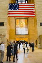 USA, New York, Manhattan, Grand Central Terminal main concourse busy with people and the Stars and Stripes flag hanging down above them.