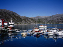 Norway, Finnmark, Nordkinnhalvoya, Fishing settlement of Dyfjord in south facing sheltered bay with fishing boats and pleasure craft moored beside wooden jetty and wind generators on skyline behind.