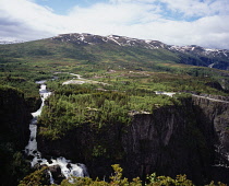 Norway, Hordaland, Sysendalen, Upper section of Voringsfossen waterfall with Halingahaugane mountain with snow patches on upper slopes against blue sky and windswept clouds behind.