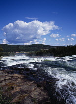 Sweden, Norrbotten, Pitealven River, View down Storforsen Rapids  25 miles north west of Alusbyn town.  Expanse of fast flowing  churning water with buildings beyond and backdrop of dense forest.  A f...
