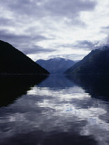 Norway, Hordaland, View east along inner Eidfjord from Bruravik.  Rippled water reflecting silvery sky with part silhouetted landscape and distant snow topped mountain.