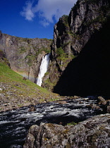 Norway, Hordaland, Mabodalen, Voringsfossen.  Waterfall crashing over steep  jagged cliff  disected by rainbow with fast flowing  rocky river in foreground.