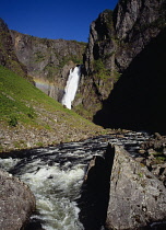 Norway, Hordaland, Mabodalen, Voringsfossen.  Waterfall crashing over steep  jagged cliff  disected by rainbow with fast flowing  rocky river in foreground.