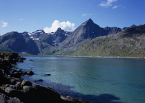 Norway, Lofoten, Flakstadpollen, Rocky shore of fjord with Stortinden Mountain peak beyond.  Snow patches lying in ravines and crevices of upper slopes and thick  white cloud at peak.