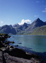 Norway, Lofoten, Flakstadpollen, Rocky shore of fjord with Stortinden Mountain peak beyond.  Snow patches lying in ravines and crevices of upper slopes and thick  white cloud at peak.
