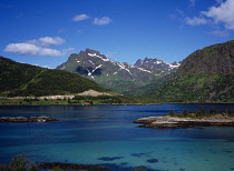 Norway, Lofoten, Indrefjorden, View across dark turquoise water of fjord with peak of Porselvtinden Mountain behind criss-crossed with lines of snow and cloud shadow.