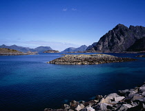 Norway, Lofoten, Vagan, Henningsvaer.  View north towards Festvagtinden Mountain and rocky islets.  Flat calm  dark turquoise water and blue sky.