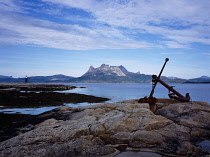 Norway, Nordland, Hamaroy, Nordneset Peninsula.  View east from Tranoy village past memorial anchor towards Tilthorn Mountain on farside of Presteid Fjorden under high  windswept cloud.