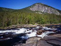 Norway, Telemark, Tessungdalen, River Austbygdaa.  Water flowing across polished granite bedrock with steep  eroded cliff face above trees behind.