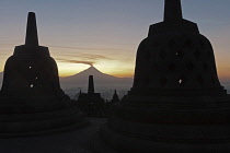 Indonesia, Java, Borobudur, Two stupas silhouetted against the sun rising behind smoking volcano, Mout Merapi.