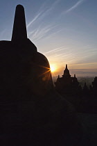 Indonesia, Java, Borobudur, Recently risen sun peeking round edge of silhouetted stupa, with volcano Mount Merapi in the background.