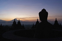 Indonesia, Java, Borobudur, Seated Buddha and stupas silhouetted just before dawn, with smoking volcano, Mount Merapi, in the distance.