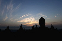 Indonesia, Java, Borobudur, Buddha and stupas silhoutted at dawn, with smoking volcano, Mount Merapi, in the background.