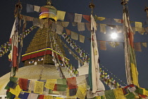 Nepal. Kathmandu, The top of the Great Stupa, viewed through flag-poles covered with Buddhist prayer flags, illuminated at night, with the moon in the background.