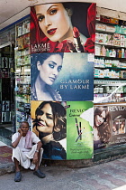 India, Maharashtra, Mumbai, Dressed in a traditional white dhoti, elderly local man sits outside a pharmacy, next to adverts for wormen's cosmetics and make up, Colaba Causeway.