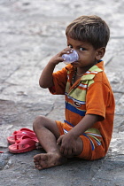 India, Maharashtra, Mumbai, Young boy drinking tea from a plastic cup while sitting on a pavement.