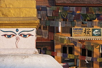 Nepal, Kathmandu, Face on mini stupa next to the Great Stupa close-up in foreground, with prayer flags and buildings in background, one with sign for Japanese restaurant.