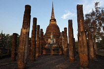 Thailand, Sukothai, Row of pillars leading up to a white seated Buddha in front of a red brick stupa, lit in the warm glow of sunset, Wat Sra Sri.