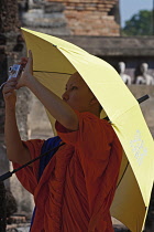 Thailand, Sukothai, Novice Buddhist monk taking a photo, shielded from the sun by an umbrella, Wat Mahathat Royal Temple.