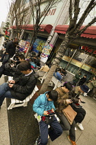 Japan, Tokyo, Akihabara, outside Yodobashi Camera mega store, men, boys and women sit on benches playing a variety of portable game consoles.
