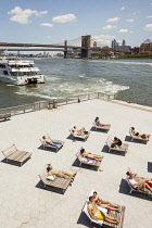 USA, New York City, Manhattan, People sunbathing beside East River, Brooklyn Bridge and South Street Seaport.
