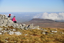 Ireland, County Fermanagh, Cuilcagh Mountain Park, Hikers sitting at the edge of a prehistoric cairn enjoying the view from the summit of Cuilcagh Mountain.