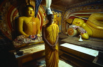Sri Lanka, Kandy, Monk standing between large seated and reclining Buddha figures in low roof interior of Nahta Revale temple.