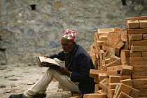 Nepal, Kathmandu Valley, Pashupatinath, Nepalese man sitting beside stack of bricks reading holy book.