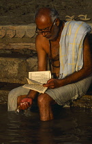 India, Uttar Pradesh, Varanasi, Man reading on steps leading to the River Ganges with his feet in the water.