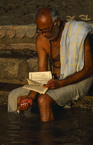 India, Uttar Pradesh, Varanasi, Man reading on steps leading to the River Ganges with his feet in the water.