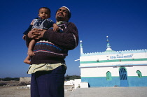 India, Gujarat, Veraval , Muslim man holding toddler with blue green and white painted mosque in background at fishing port on the south coast of Saurashtra.