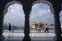 India, Punjab, Amritsar, Golden Temple, Visitors on black and white marble walkway beside sacred pool with view to temple beyond framed by arches of covered colonnade.