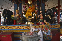 Malaysia, Penang, Georgetown, Wat Chayamangkalaram, Interior with woman placing offerings on altar in front of seated Buddha covered in gold leaf, Incense burning in foreground.